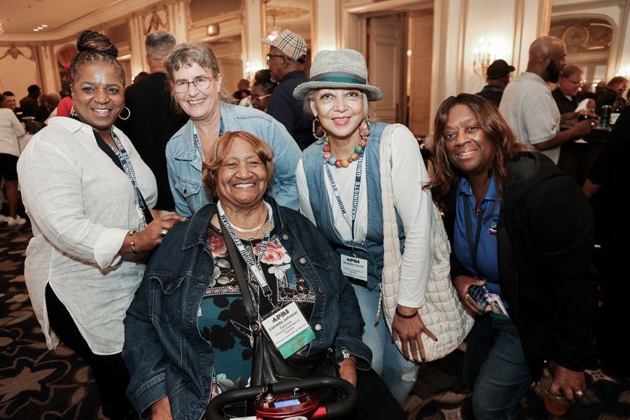 Group of women smiling at an event. One uses a mobility scooter. Formal setting with background people.