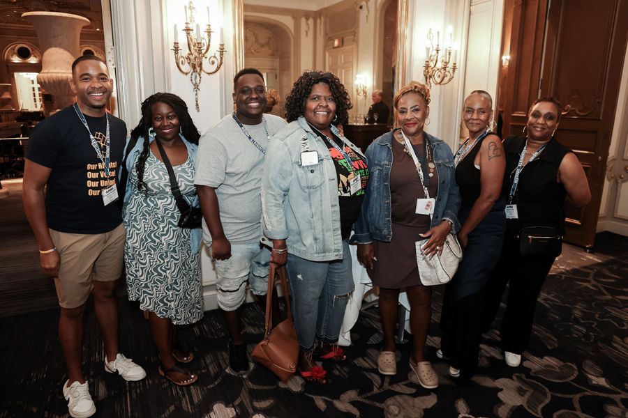 Group of eight people posing in an ornate room. People are smiling, wearing casual clothes and lanyards.