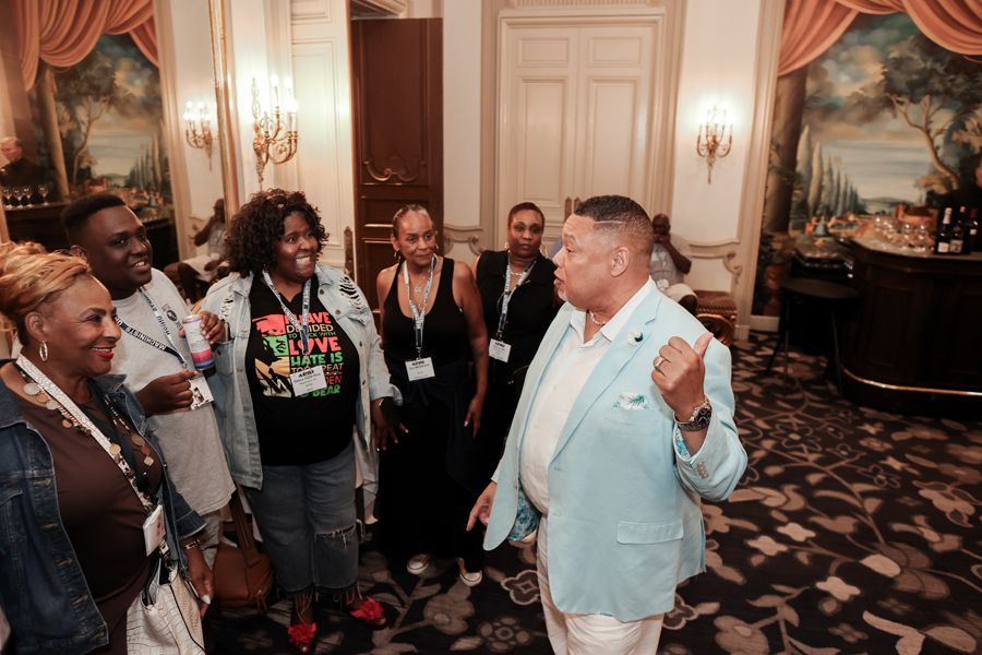 A group of people socialize in an ornate room; a man in a blue suit speaks animatedly.