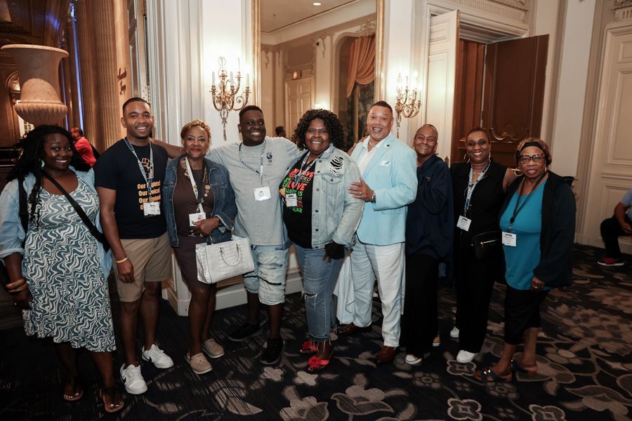 Group of diverse people smiling, posing for photo in ornate hall with chandeliers and mirrors.