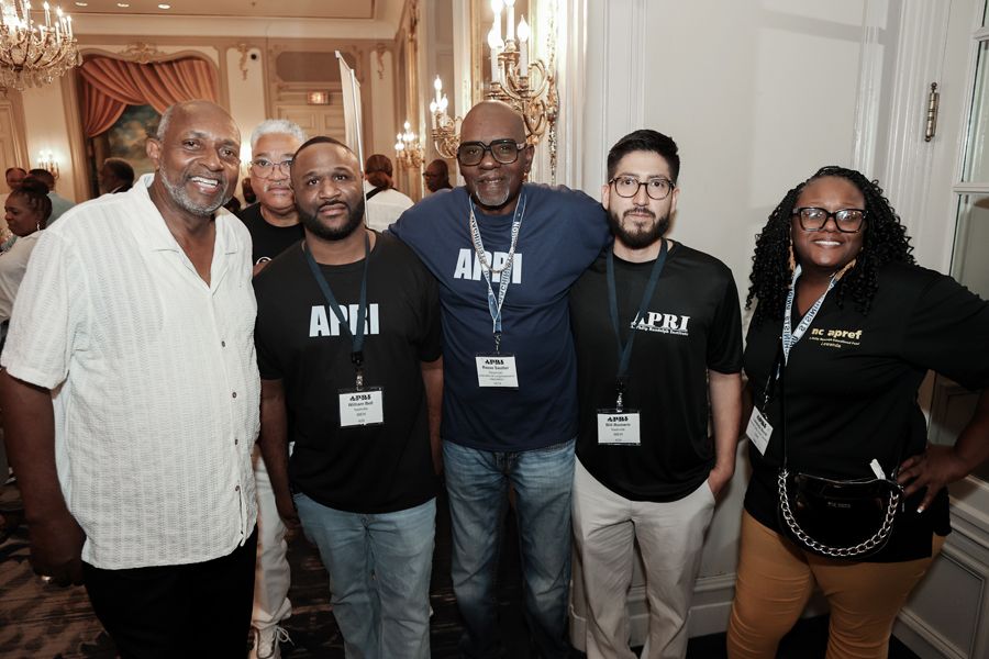 Group of people smiling in an ornate room. People are wearing lanyards and black shirts with logos.