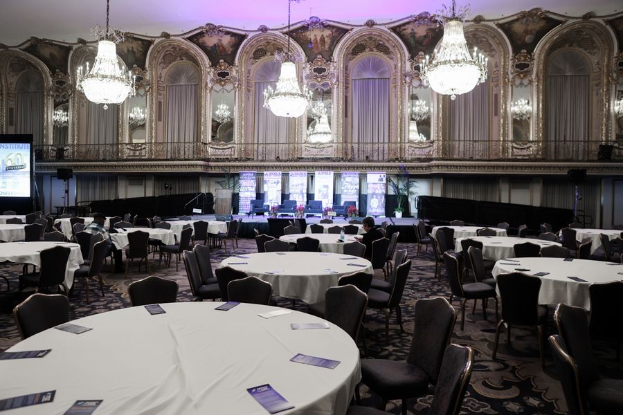 Ballroom with round tables set for an event, stage at the back, chandeliers, and ornate architectural details.