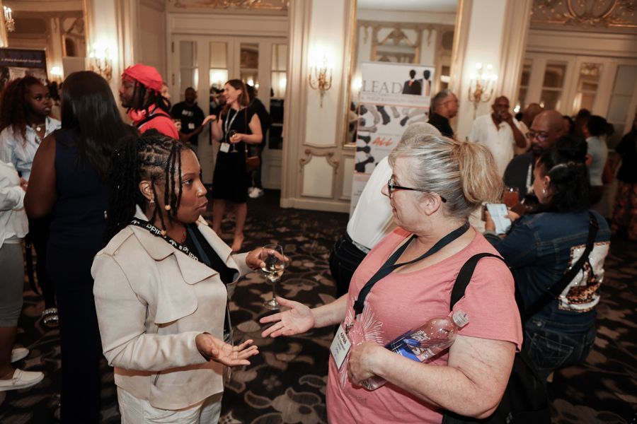 People networking at an event in a large hall with ornate doors. Two women are talking and holding drinks.