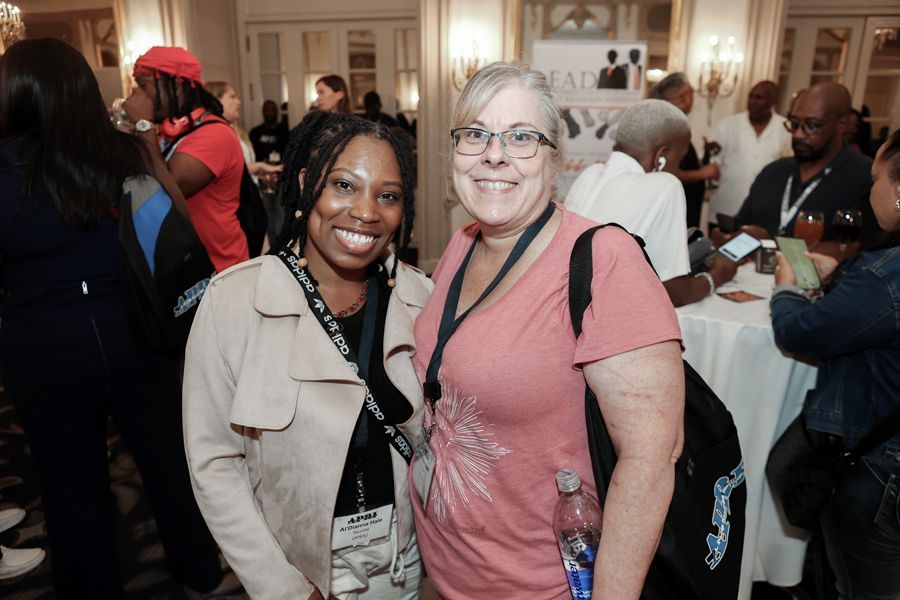 Two women smile, posing together at an event; one in a tan coat, the other in a pink shirt.