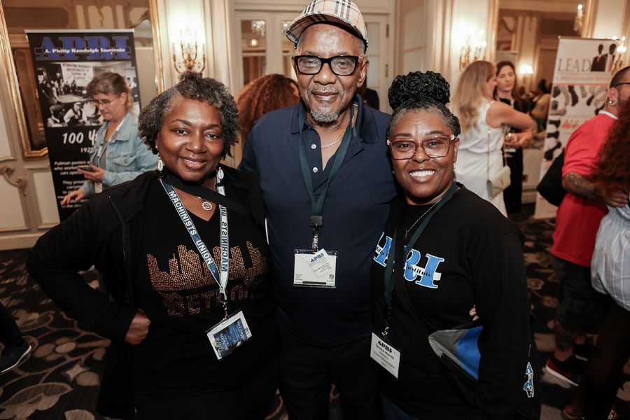 Three people smiling in a room; a man in a hat and glasses flanked by two women, all wearing name tags.