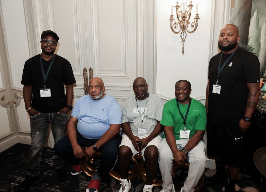 Five men posing, some seated. In a well-lit room with decorative sconce and wall paneling.