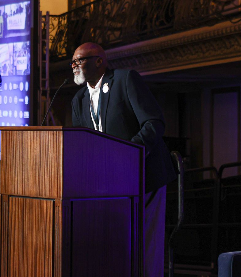 Man speaking at a podium, wearing a blazer and white shirt, in a formal setting.