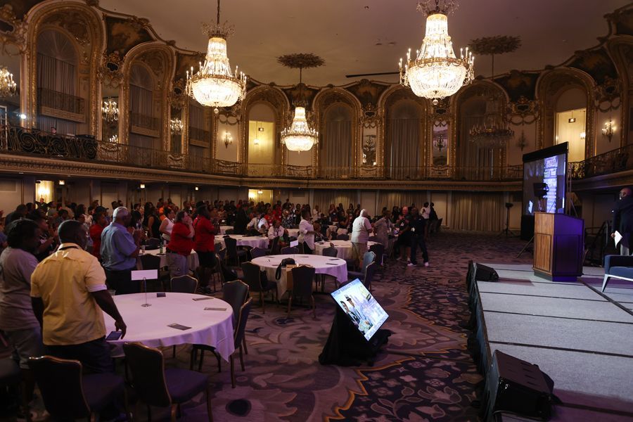 People gathered in a ballroom, attending a conference. Tables and a stage are set up. Ornate chandeliers hang.