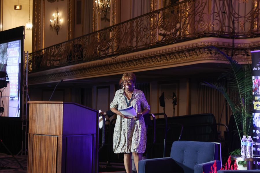 President Clayola Brown walking to podium presenting in a ballroom with ornate balcony in background.
