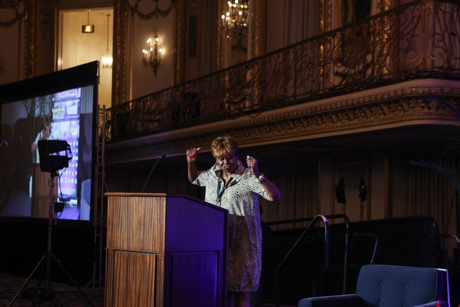 President Clayola Brown celebrating at a podium, gesturing with hands. A screen displays an image in a grand hall.