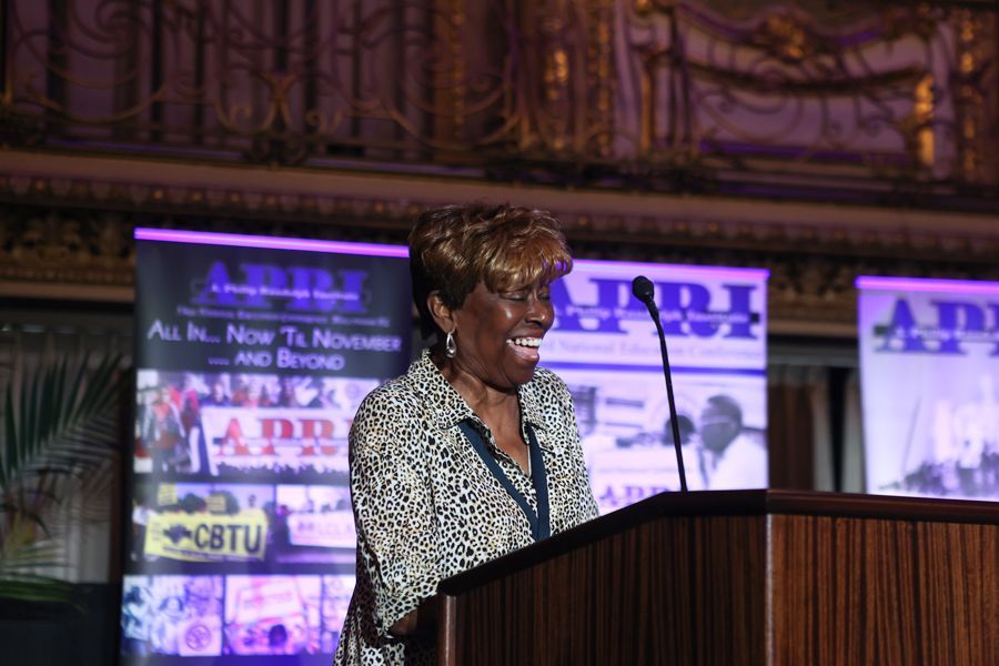 Woman speaking at a podium, smiling. Backdrop features 