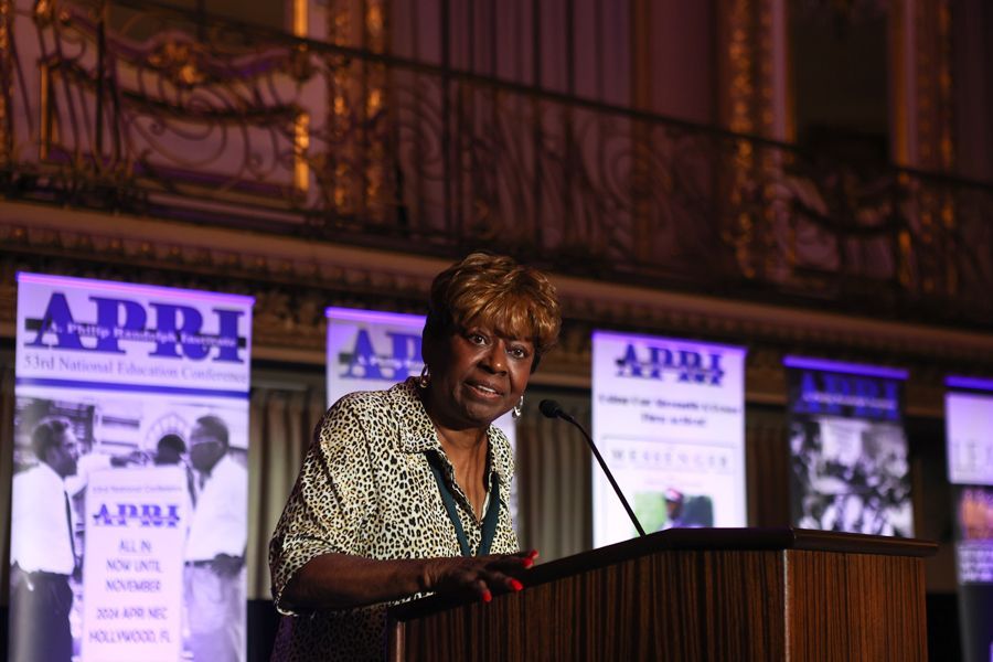 Woman speaking at podium, at conference. Beige top, floral print. Banners with logos in background.