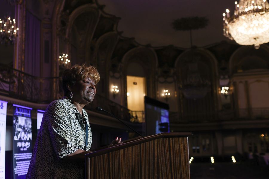 Woman speaking at a podium in a grand hall with chandeliers; she's smiling and wearing a patterned jacket.