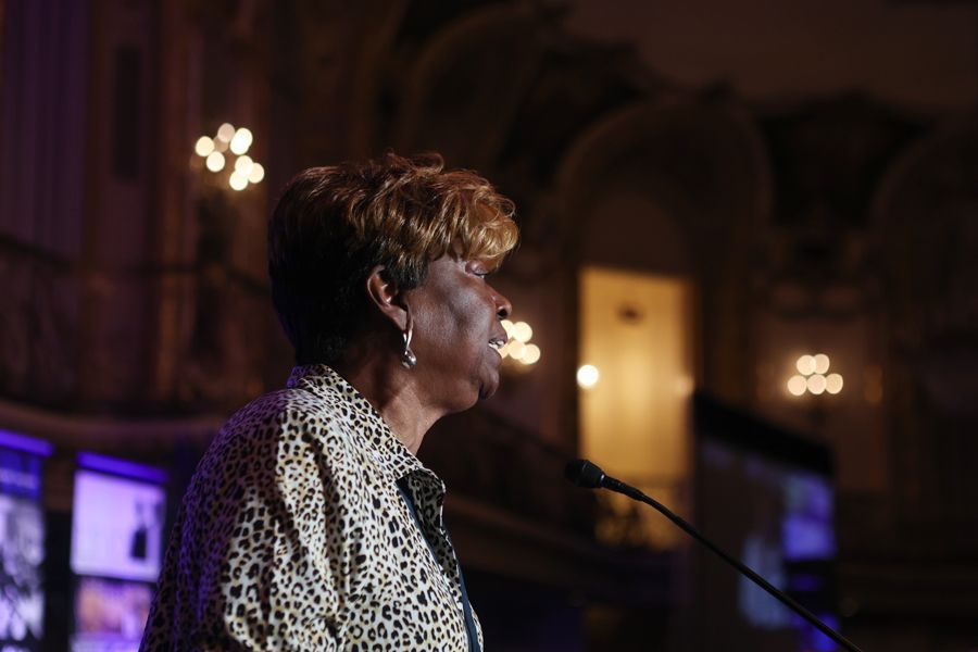 Clayola speaking at a podium in a low lit auditorium, wearing a patterned jacket.