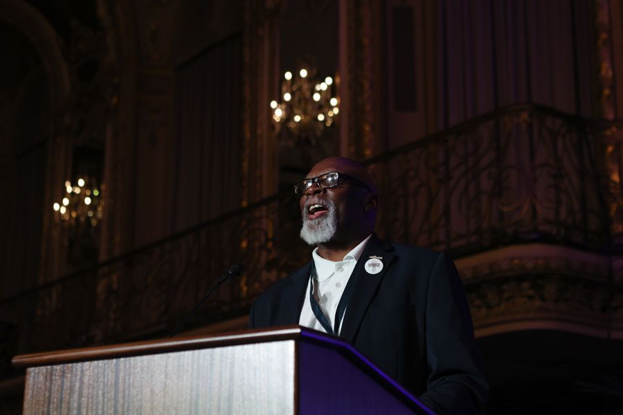 Man speaking at a podium in a dimly lit hall, wearing glasses and a blazer.