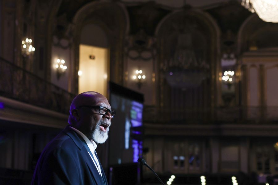 Man speaking at a podium in a formal hall with ornate architecture, lights, and a screen.