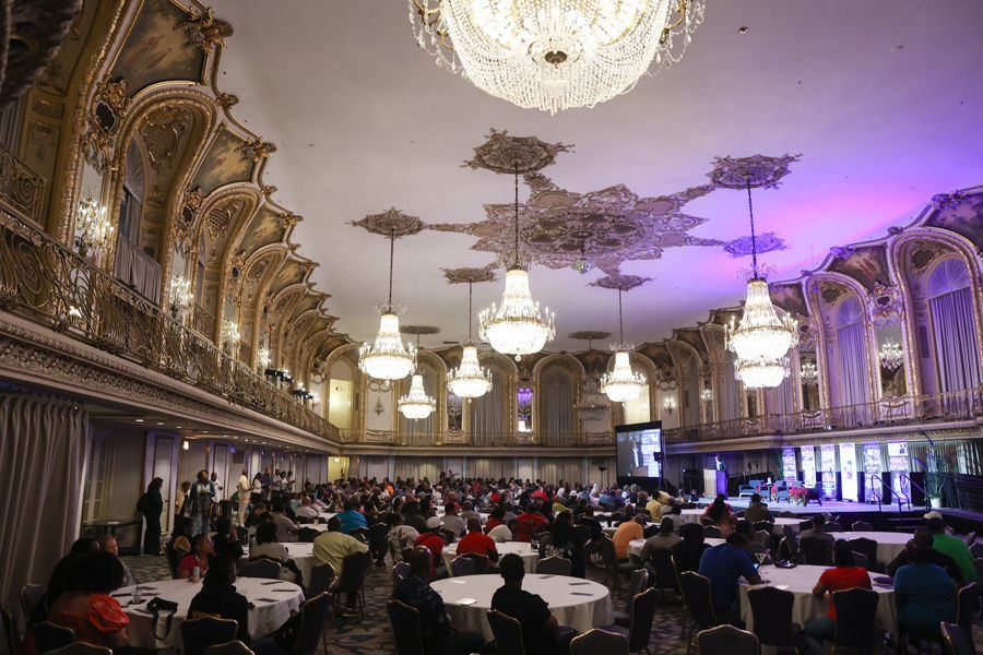 Large ornate ballroom with many people at round tables; chandeliers hang from ceiling.
