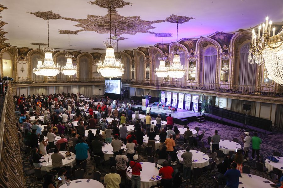Large event in ornate ballroom: many people stand and sit at tables facing a stage with a screen.