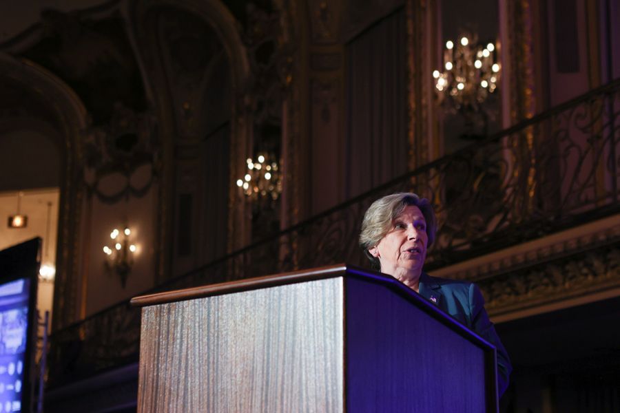 Woman speaking at a podium in a grand ballroom, with chandeliers and ornate details.
