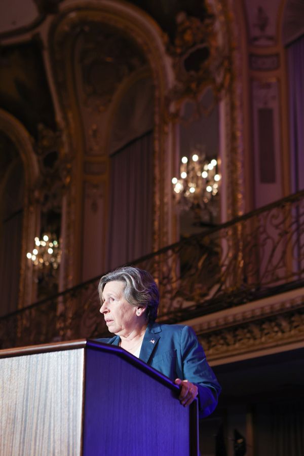 A woman speaking at a podium in an ornate ballroom, wearing a suit, looking to the side.
