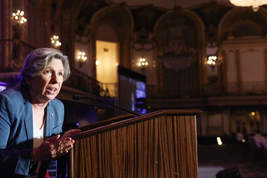 Woman in blue blazer speaks at a podium in an ornate room; she has a focused expression.