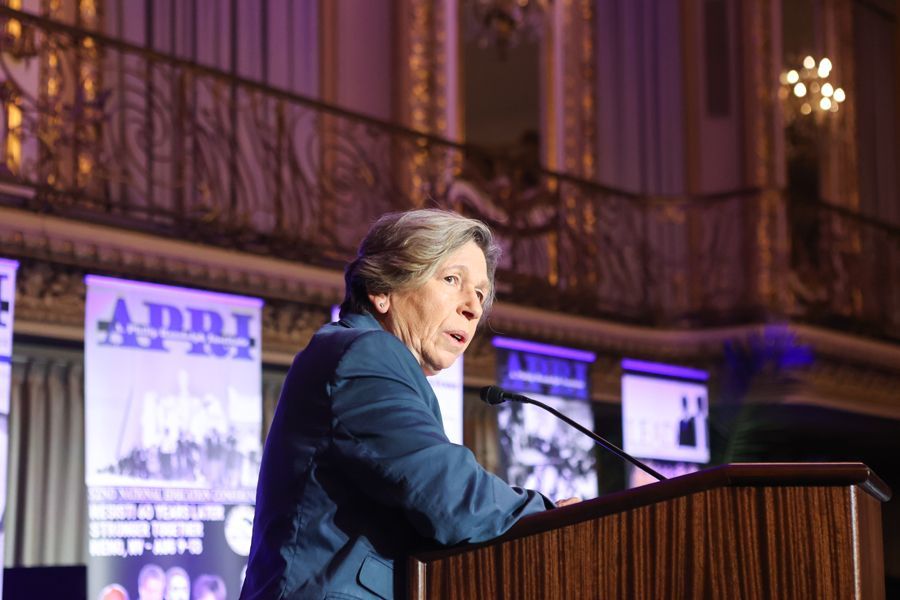 Woman speaking at a podium, wearing a blue blazer, inside a decorated hall with banners.