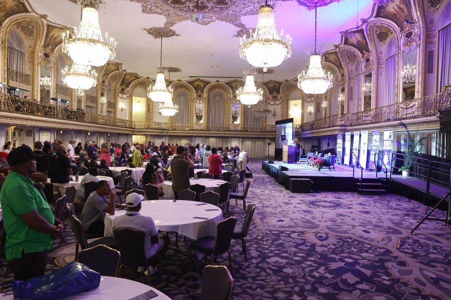 Large ballroom with chandeliers and people attending an event with a stage.