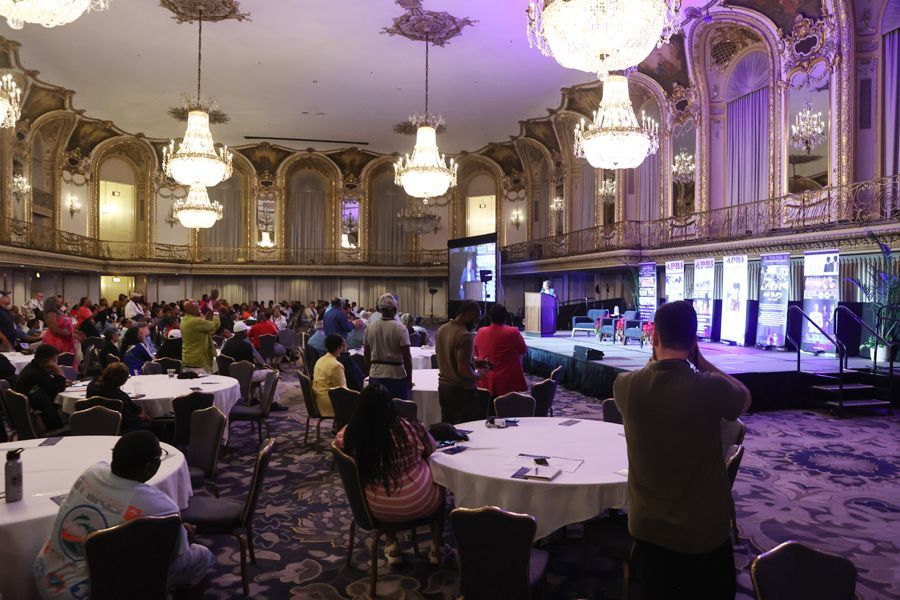 A large ornate ballroom with people seated at tables, a stage with speakers, and chandeliers.