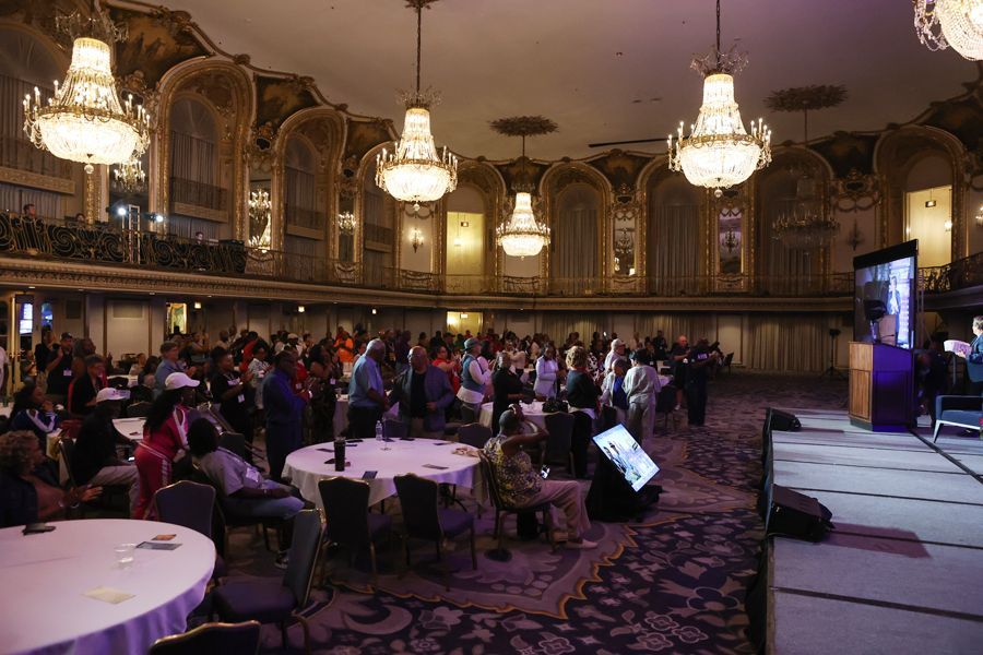 Audience in a ornate ballroom watches a presentation on a stage, with chandeliers above.
