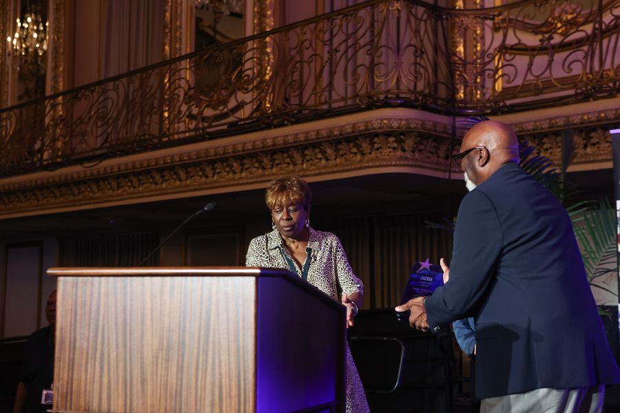 Woman at podium in ornate hall, man approaching. Bronze, gold accents.