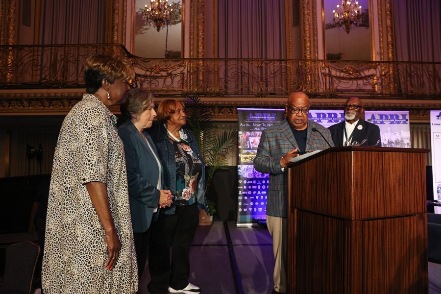 People at a podium with a speaker; others stand to his side at an event in a decorative room.