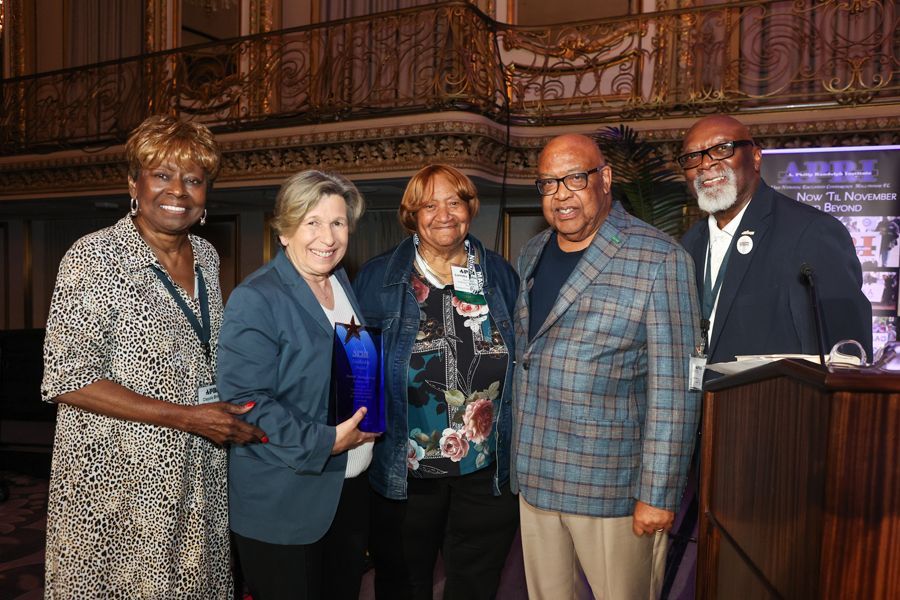 Five people at a podium, posing for a photo. They are smiling. Interior with ornate balcony.