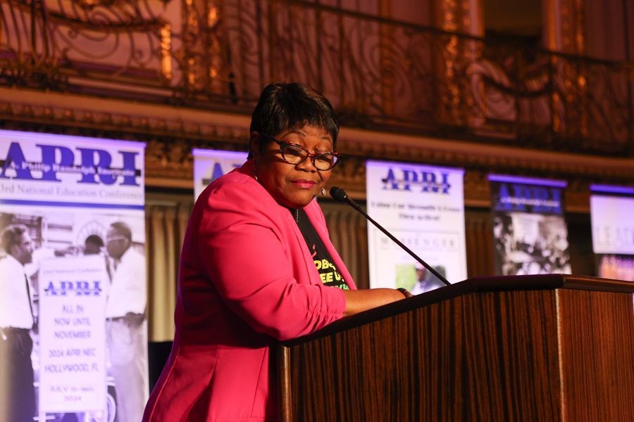 Woman in pink blazer speaks at a podium with event banners in a grand hall.