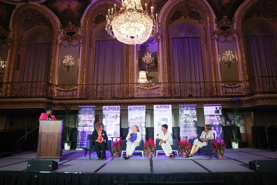 Panel of women on stage at an event. They are seated. Ornate ballroom setting, purple banners.