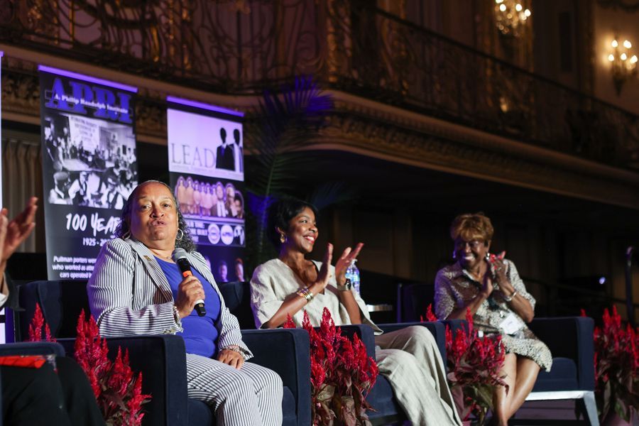 Three women on stage at an event, seated on a sofa, clapping. Purple and black banners in background.
