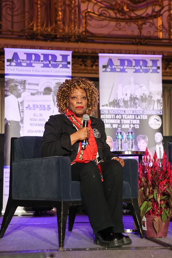 Famous Actress in black suit with red scarf, seated on stage, holding a microphone, with APRI banners in the background.