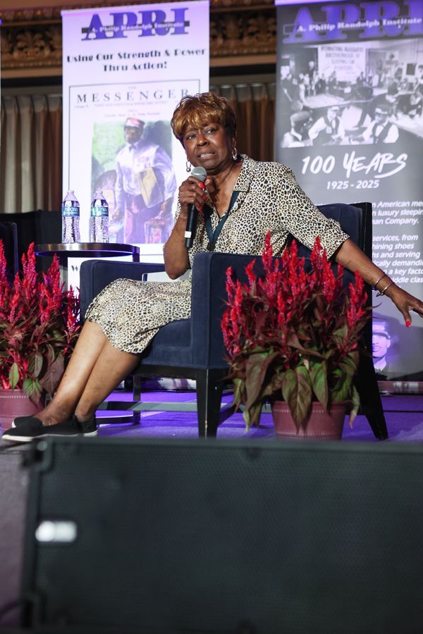 Clayola Brown speaks into a microphone, seated on stage with floral accents.