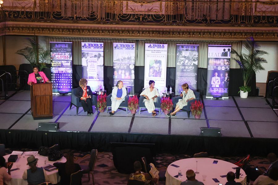 Panel discussion on a stage. Speakers in front of banners, seated in chairs, speaking to an audience.