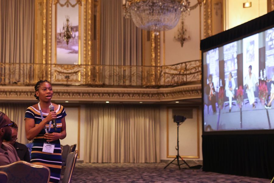 young woman participating at a conference, standing in front of a screen, a large chandelier hangs above.