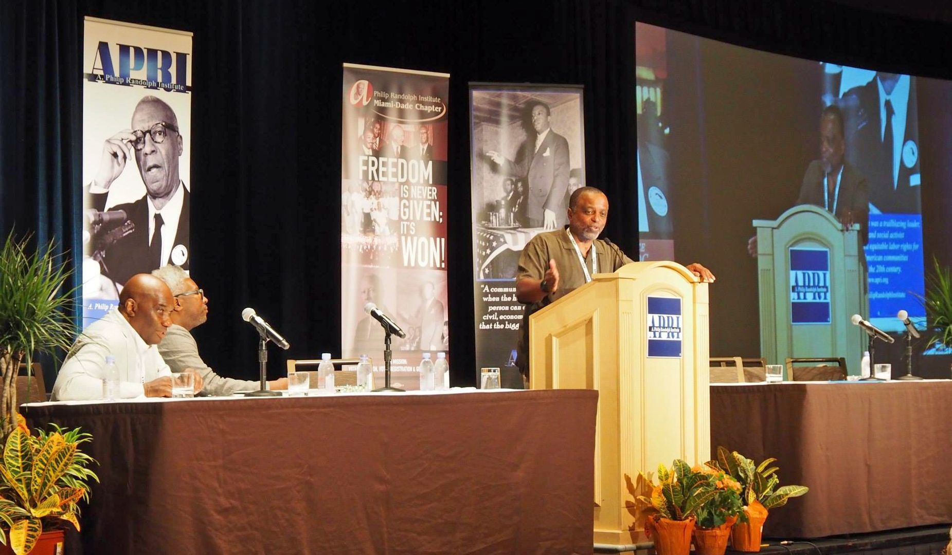 A man stands at a podium in front of a screen that says april