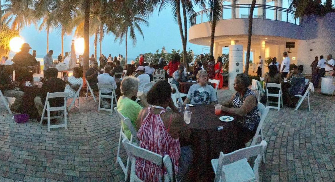 A group of people are sitting at tables outside in front of palm trees.