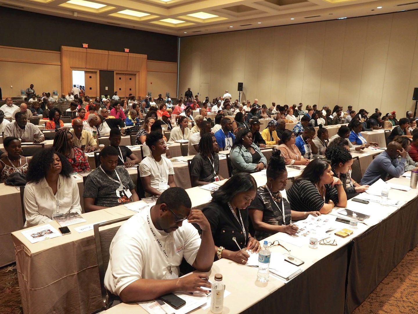 A large group of people are sitting at long tables in a conference room.