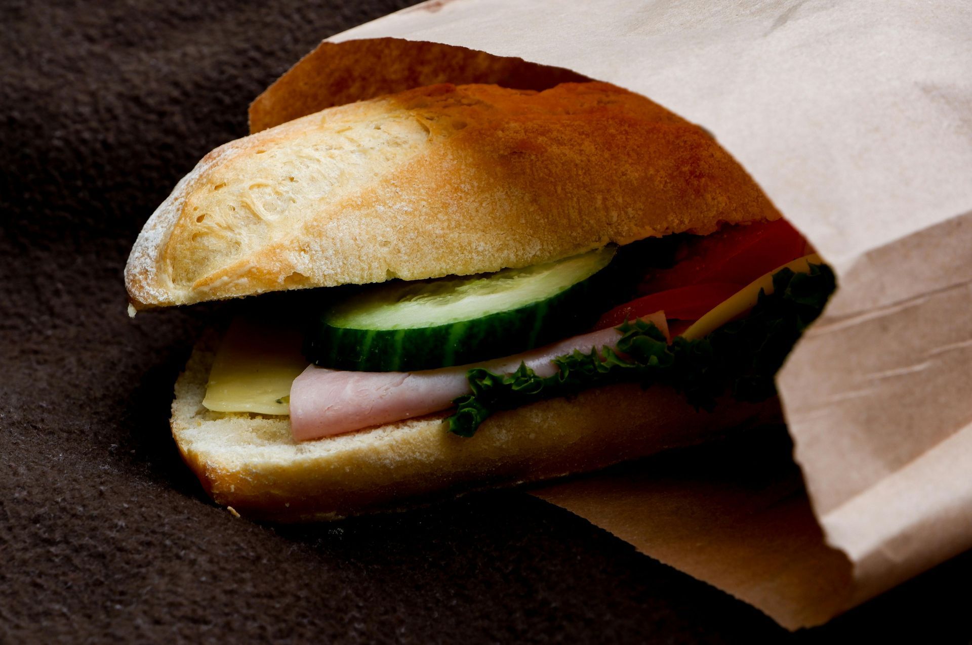 Una mano che regge un panino farcito con verdure, formaggio e salumi su carta bianca, accanto a un piatto bianco.