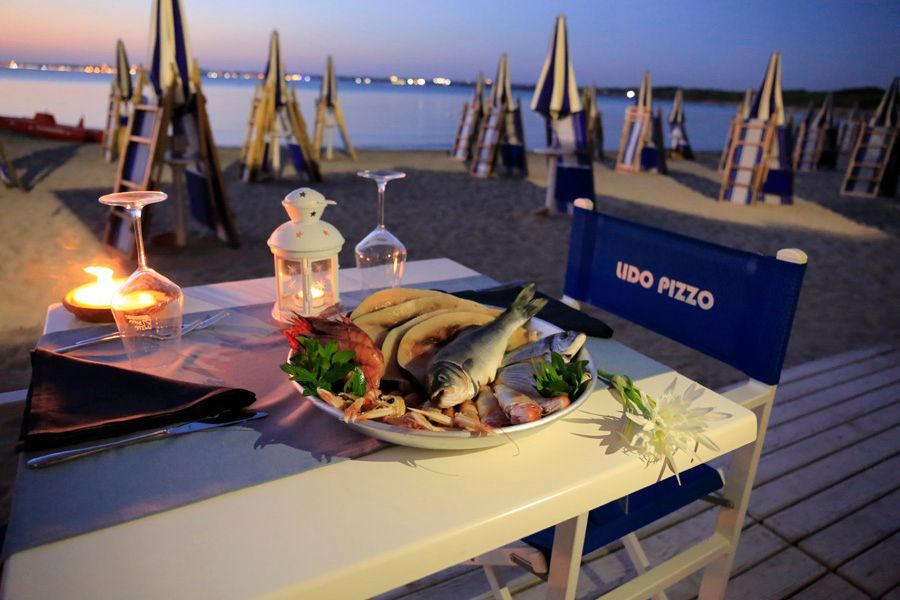 Tavolo da pranzo in riva al mare con pesce alla griglia, insalata e bevande al tramonto.