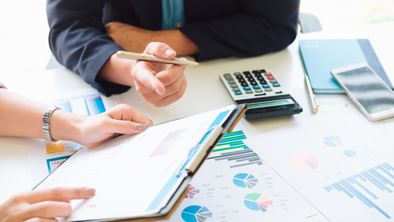 Two professionals review financial charts and data on a document at a desk with a calculator and smartphone nearby.