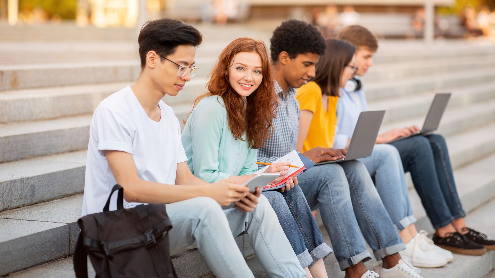 A group of students sit on outdoor stone steps, using laptops and notebooks while studying together.