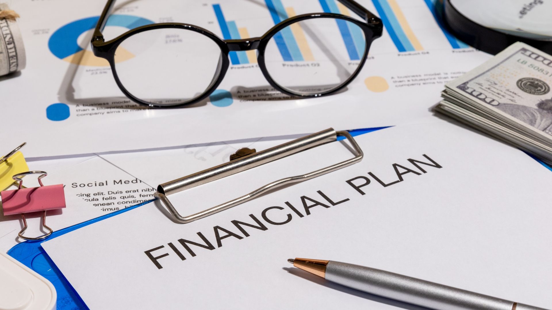 A Financial Plan document with a pen, eyeglasses, and a stack of cash on a desk with financial charts.
