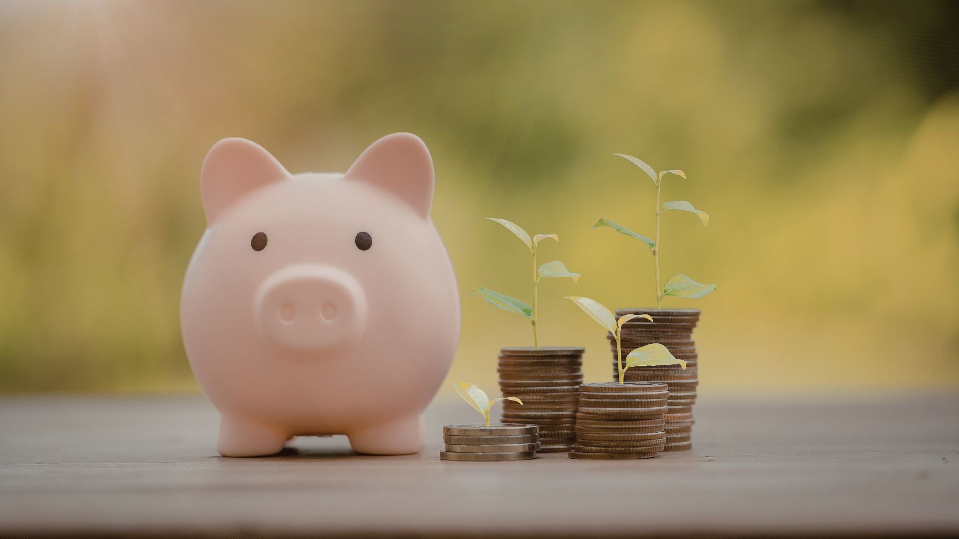A light pink piggy bank sits next to three small stacks of coins, each topped with a small green plant, on a wooden surface.