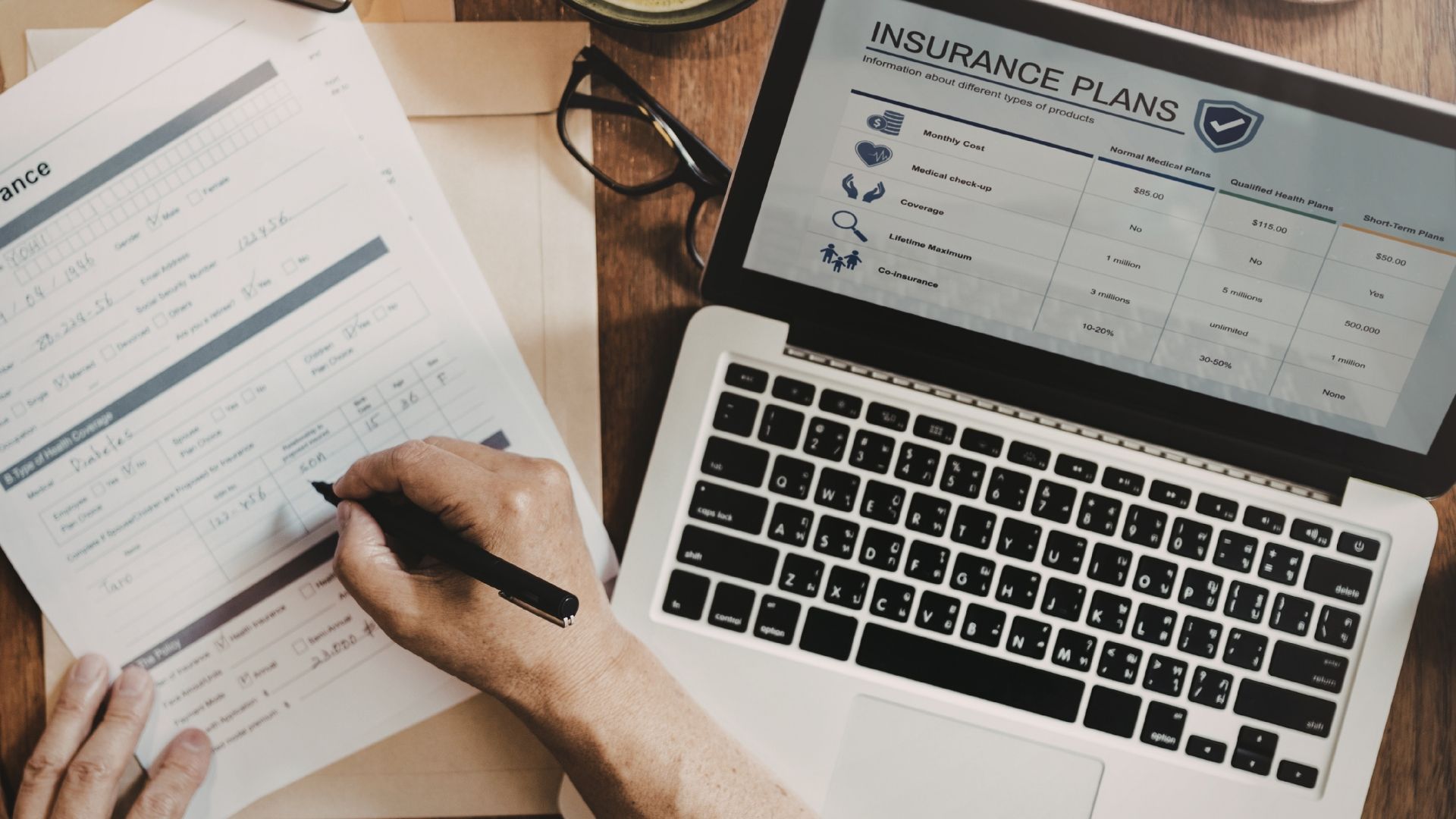 Hands filling out a form next to a laptop displaying an insurance plan comparison table on a wooden desk.