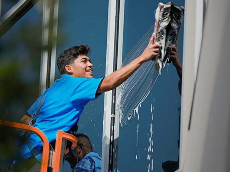 A person in a blue shirt standing in an orange lift, cleaning a tall glass building with a squeegee.